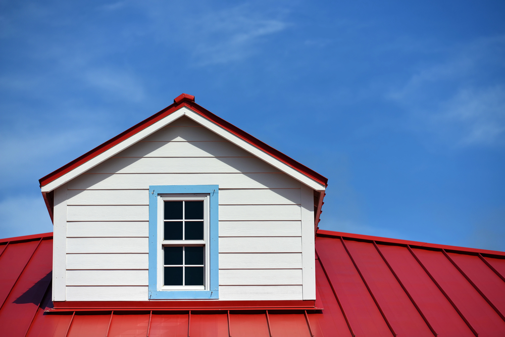 White dormer with a blue-trimmed window on a red metal roof against a clear blue sky.