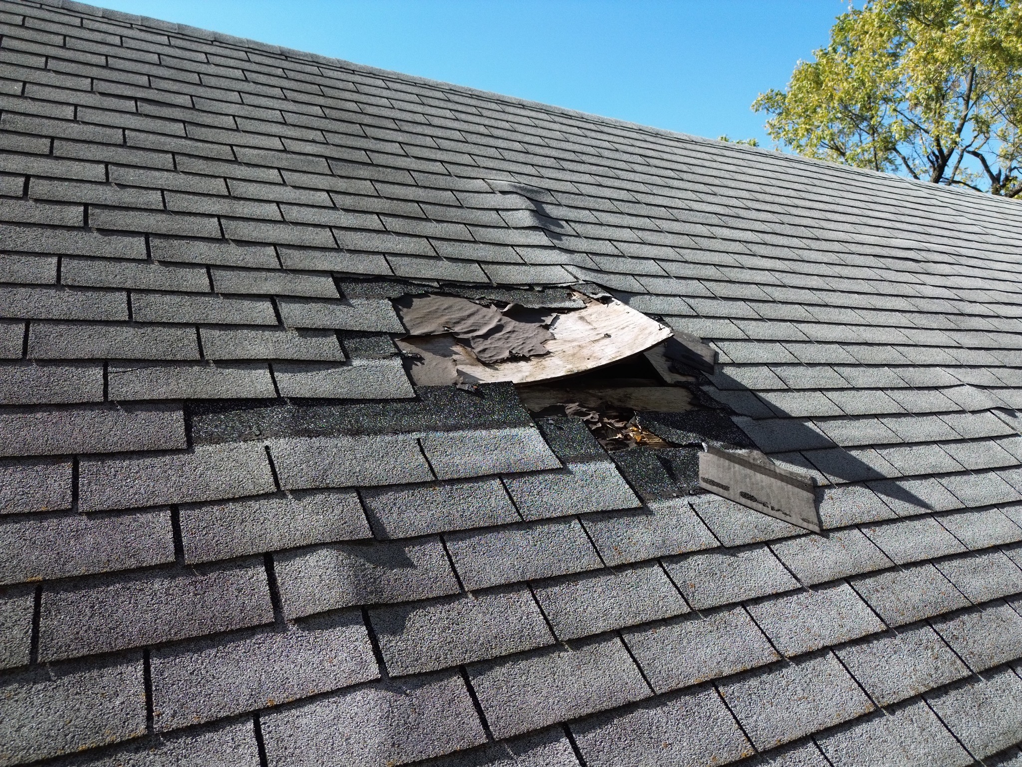 A house roof with a hole in the shingles, exposing damaged wood underneath against a clear blue sky.