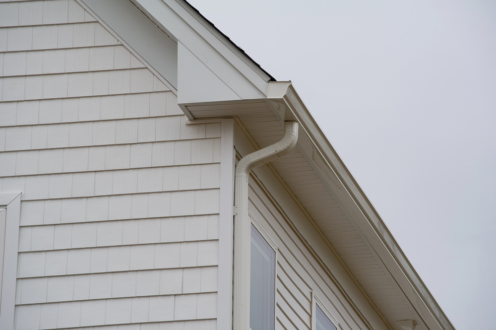 Close-up of a house exterior showing white siding, windows, a roof edge, and a white downspout under an overcast sky.