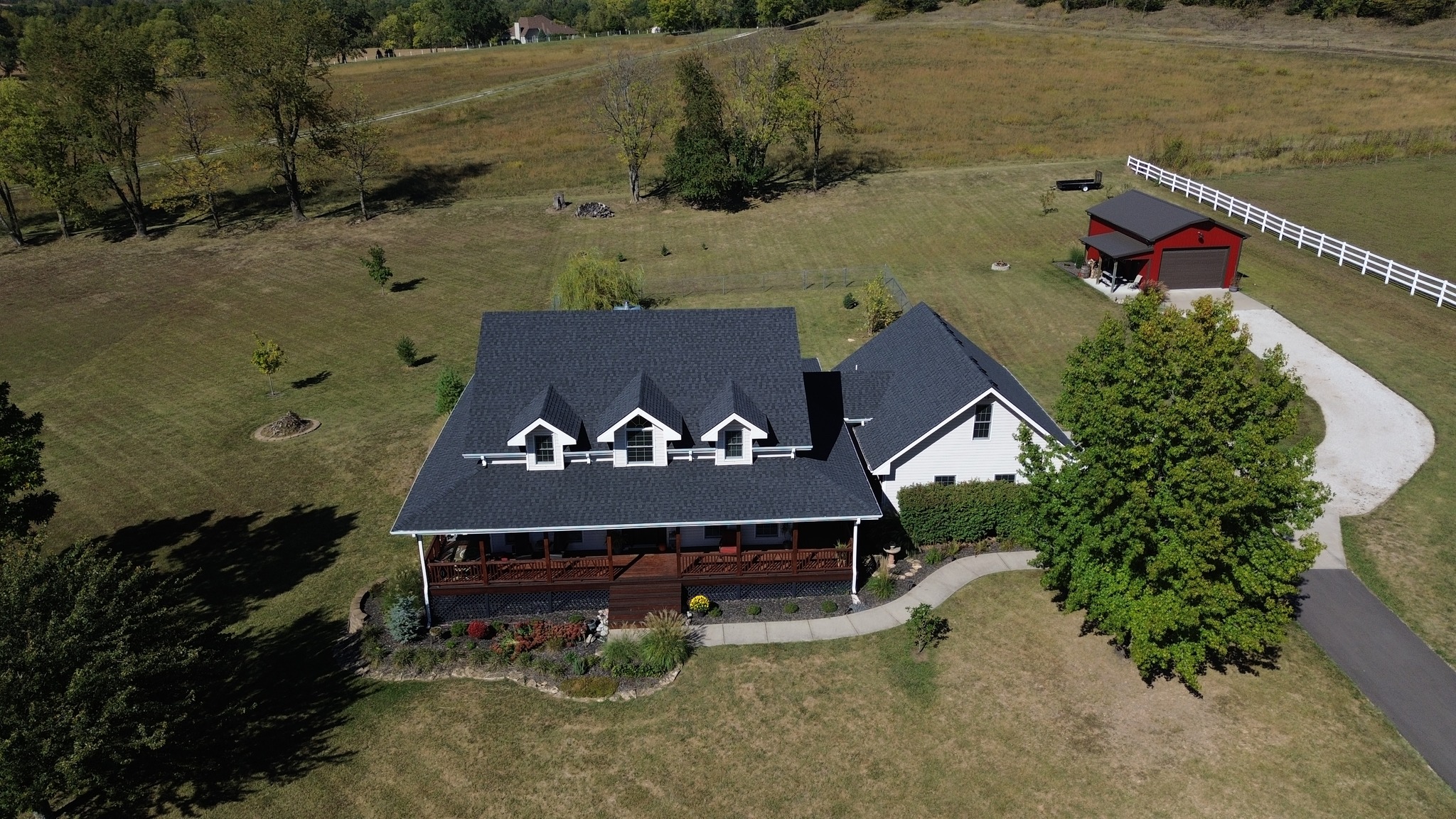 Aerial view of a house with a covered porch, surrounded by lawn and trees, with a small red outbuilding and a curved driveway in a rural setting.