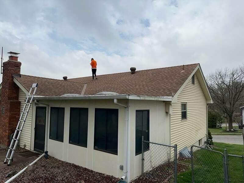 A person in an orange shirt stands on the roof of a beige house with a ladder leaning against the side.